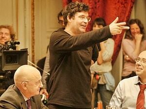 Pierre Schoeller, au centre de la photo, regarde vers la droite, la main tendue vers la même direction. Il est en pleine conversation avec deux acteurs, l'un à droite de l'image et l'autre à gauche.