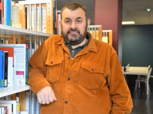 Rémi Lefebvre photographié de face, en plan rapproché dans la bibliothèque universitaire. Il regarde l'objectif et son bras droit est accoudé sur le rayon d'une étagère de livres.